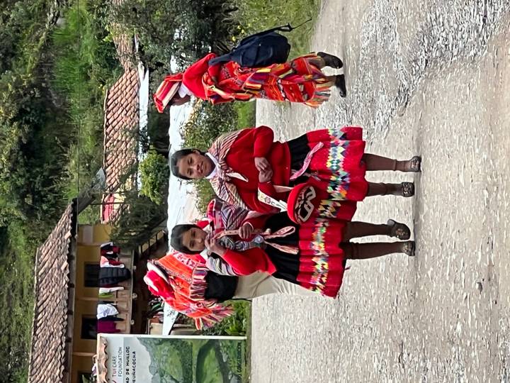 Two young girls in bright traditional Andean clothing walk along a rural road with villagers in the background