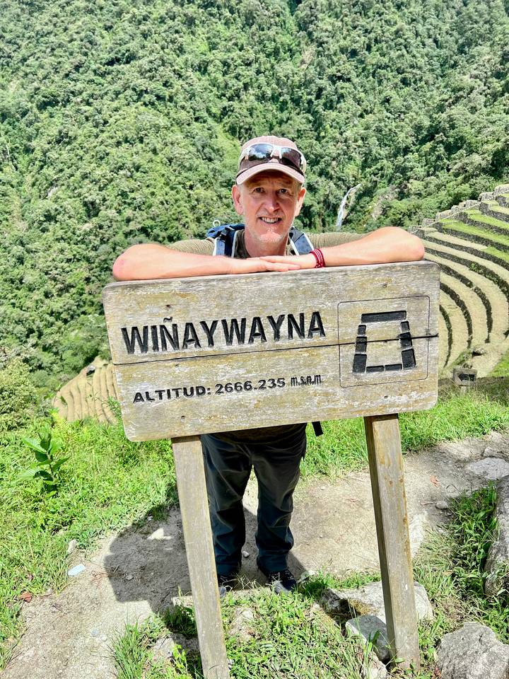 Traveler leaning on a weathered wooden sign reading 'Wiñaywayna' with terraced mountains behind