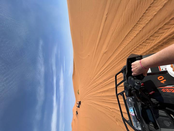 Rider steering a quad bike across vast orange desert dunes under a clear blue sky