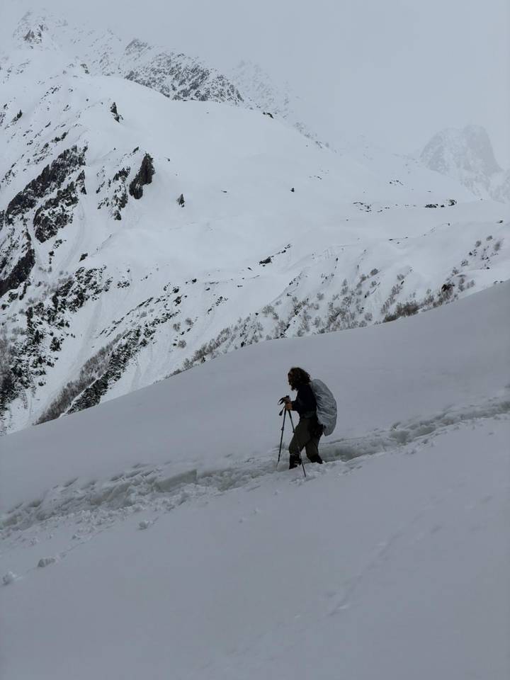 A trekker with poles ascends a snowy mountainside beneath towering white-clad peaks.