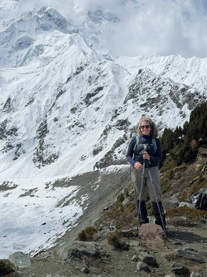 A smiling hiker poses with trekking poles against a backdrop of steep snow-covered slopes and conifers.