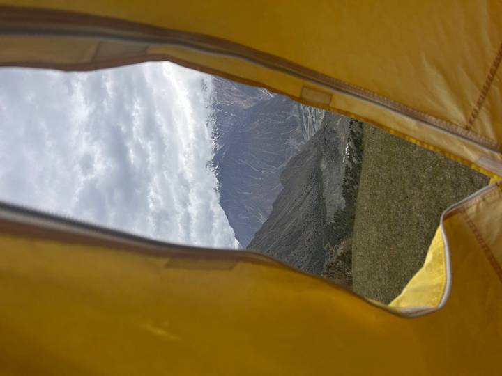 Mountain peaks dusted with snow are framed by the open flap of a yellow tent on a grassy plateau.
