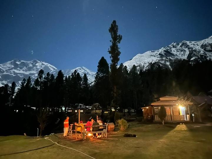 Under a starry moonlit sky, travellers sit around a campfire with snow-capped peaks towering above dark pines.
