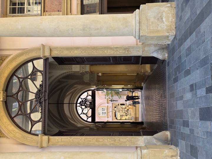 Historic archway leading to a courtyard where two people admire a baroque fountain sculpture.