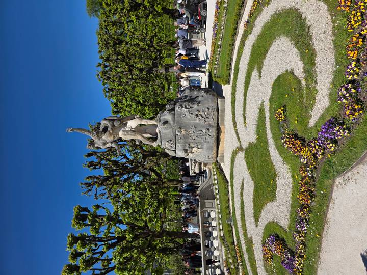 Stone fountain statue in manicured garden maze with crowds touring on a bright spring day.