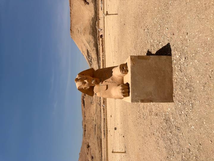 Small sphinx statue rests on a stone plinth in a vast sandy archaeological area under a blue sky.