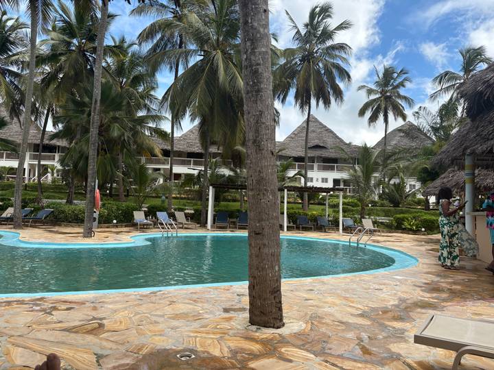 Resort swimming pool bordered by palm trees and thatched buildings on a sunny tropical day