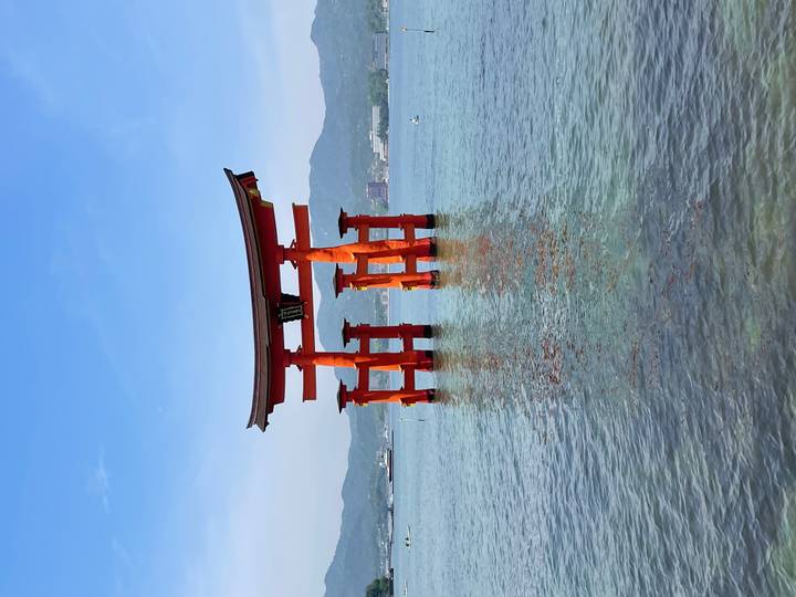 Vibrant red torii gate of Itsukushima Shrine standing in calm blue water with distant hills under a clear sky
