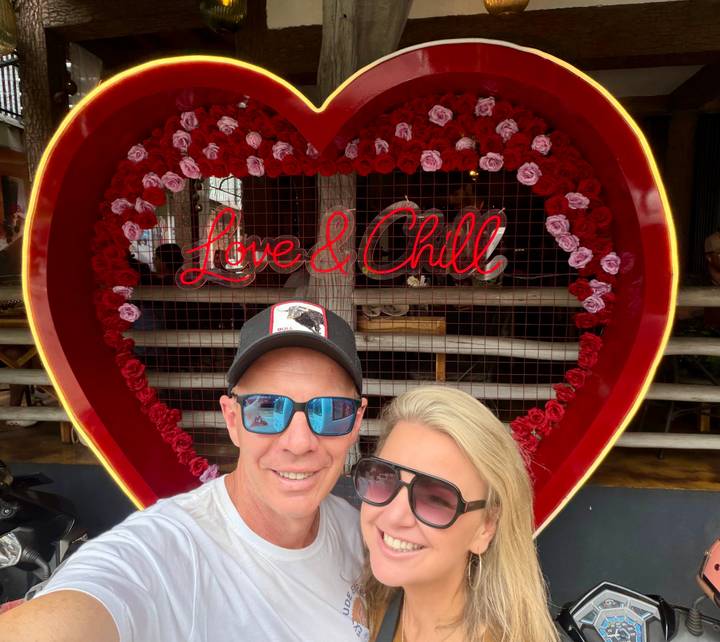 Couple takes a selfie in front of a heart-shaped floral frame with neon words 'Love & Chill'.