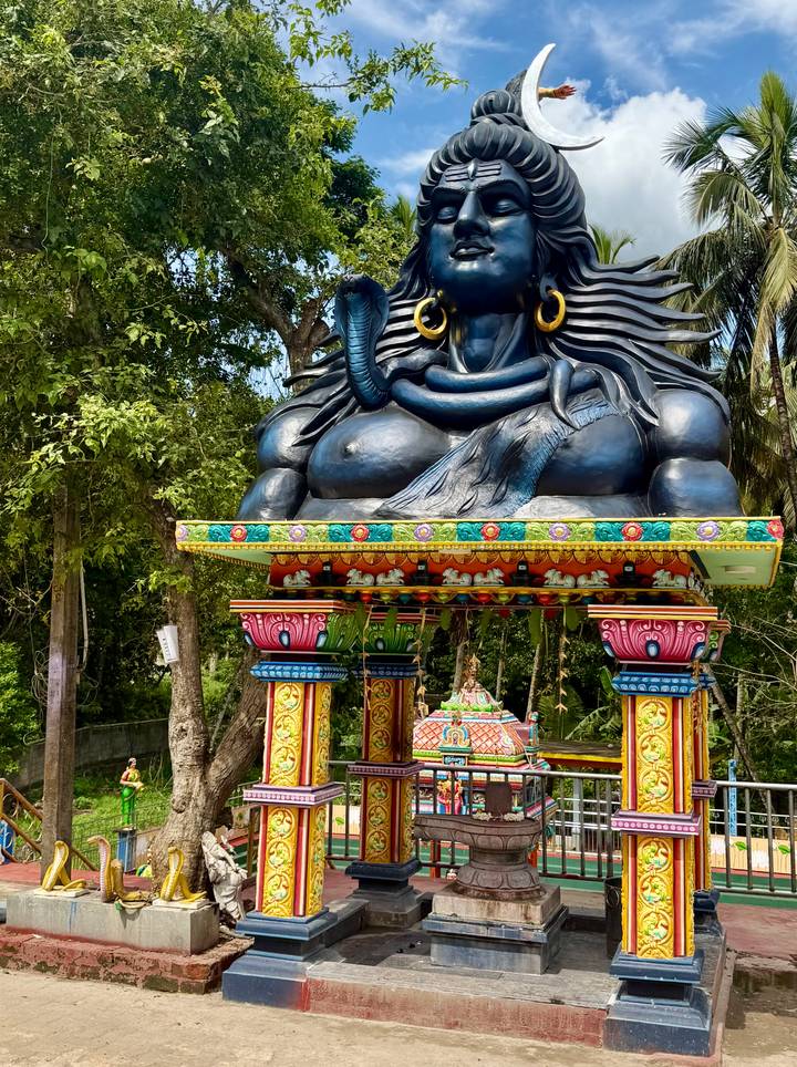 Vibrant multicoloured shrine featuring a large seated Shiva figure surrounded by jungle greenery.