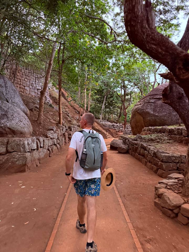 Man with backpack walks a red-earth path through ancient stone terraces toward steep forested steps.