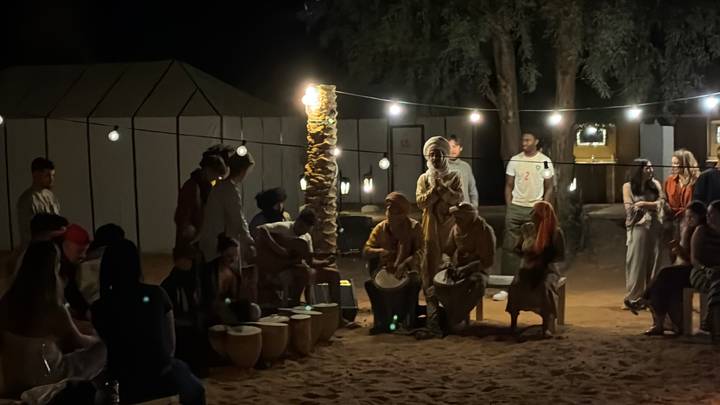 Nighttime desert camp scene with travelers drumming and string lights illuminating the sand