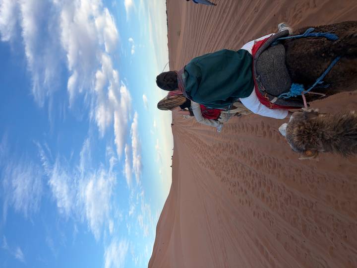 Travelers ride camels in single file across vast red sand dunes under blue sky