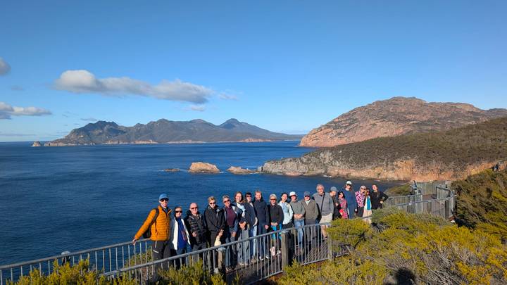 A tour group poses on a coastal lookout railing with sweeping views of rugged mountains and blue sea.