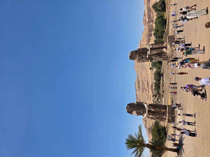 Visitors gather before the towering Colossi of Memnon under a vast blue sky.