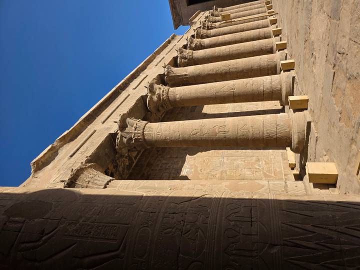 Exterior view of sandstone columns with hieroglyphs at an ancient Egyptian temple against blue sky.