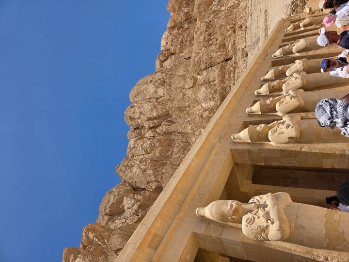 Colonnade of statues of Hatshepsut with desert cliff backdrop at temple complex, under clear blue sky with tourists below.