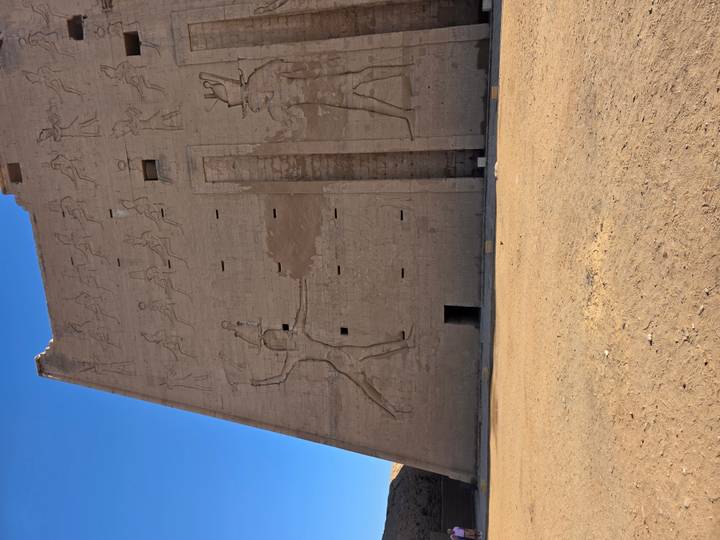 Massive blank temple pylon wall with carved figures and empty sandy forecourt under blue sky.