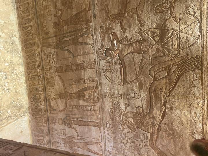Temple wall relief showing chariot-riding pharaoh aiming bow among hieroglyphic panels.