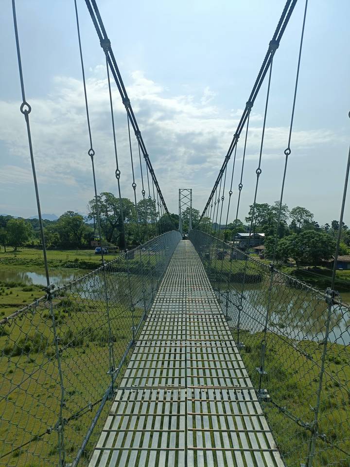 Metal suspension bridge stretching over a reflective river with verdant countryside beyond