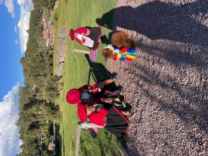 Women in traditional Andean dress sit beside a baby alpaca wearing a rainbow scarf