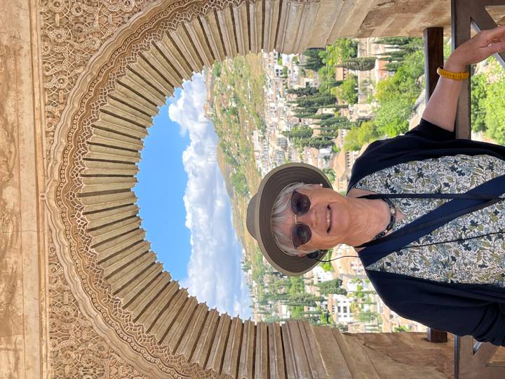 Smiling older woman posing beneath an ornate Moorish archway with Granada’s hills and cityscape framed behind her.