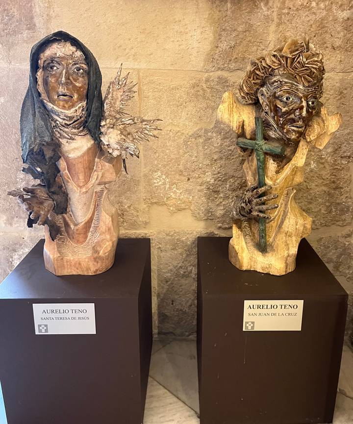 Two modern religious-themed wooden sculptures displayed on dark plinths against a stone wall inside a gallery.