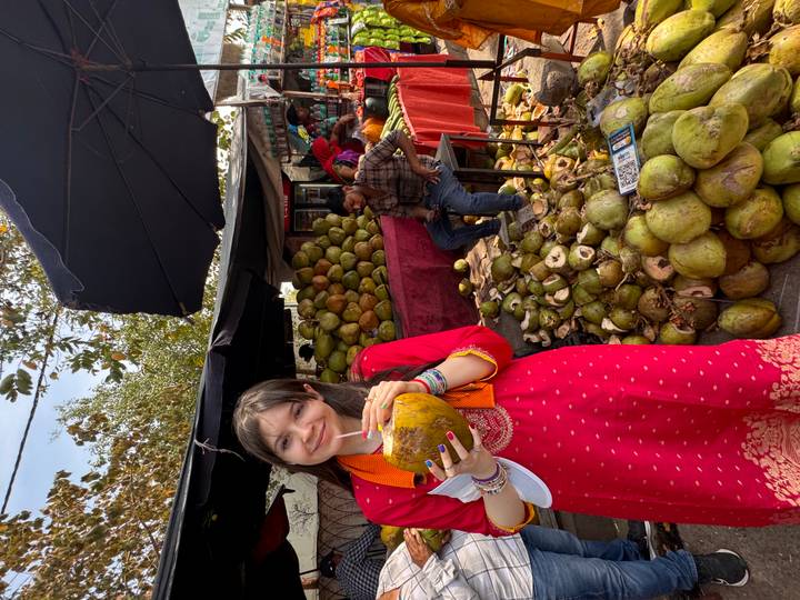 Traveller in traditional dress drinks fresh coconut at a bustling fruit stall.