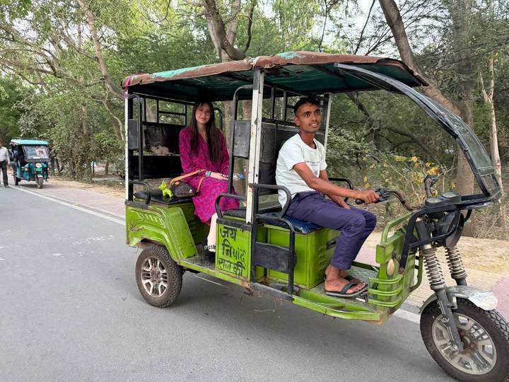 Passenger rides in a green e-rickshaw along a tree-lined Indian roadway.