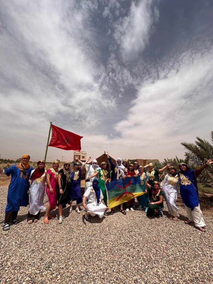 Large tour group in colourful traditional dress posing with Moroccan flags in an arid village setting