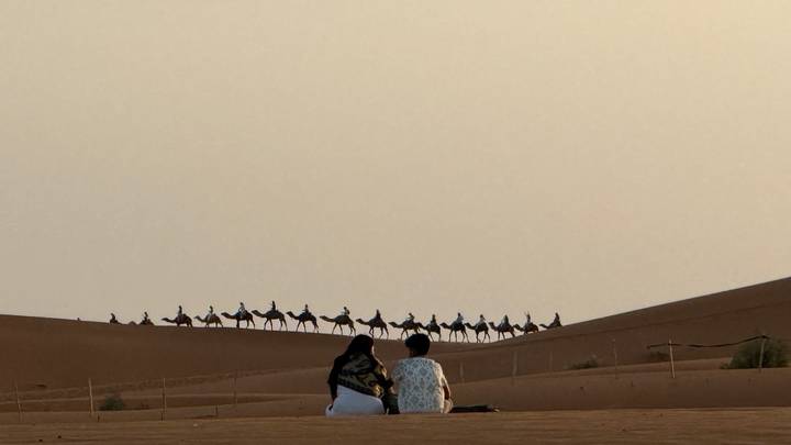 Two people sitting on dune watching long caravan of camels crossing desert ridge at dusk.