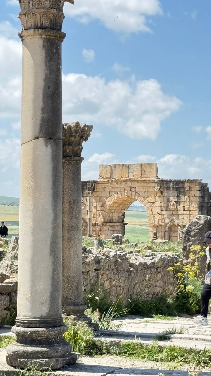 Stone arch and columns of ancient Roman ruins set against green rolling hills and blue sky.