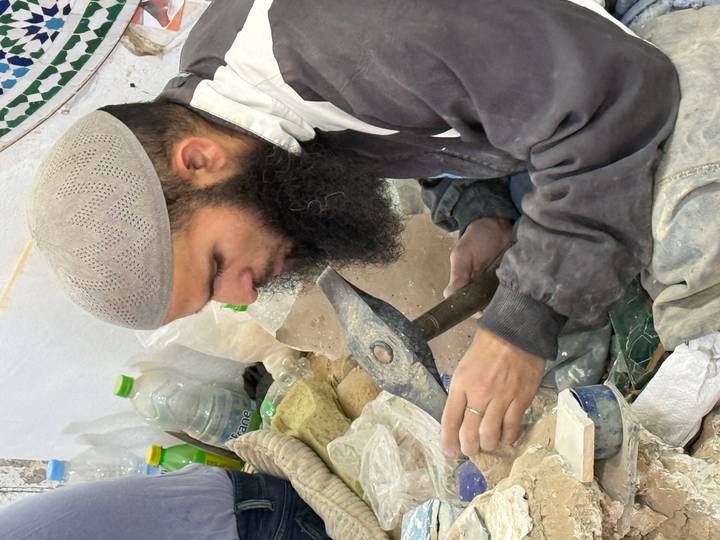 Craftsman with long beard carving stone with hammer in workshop surrounded by tools.