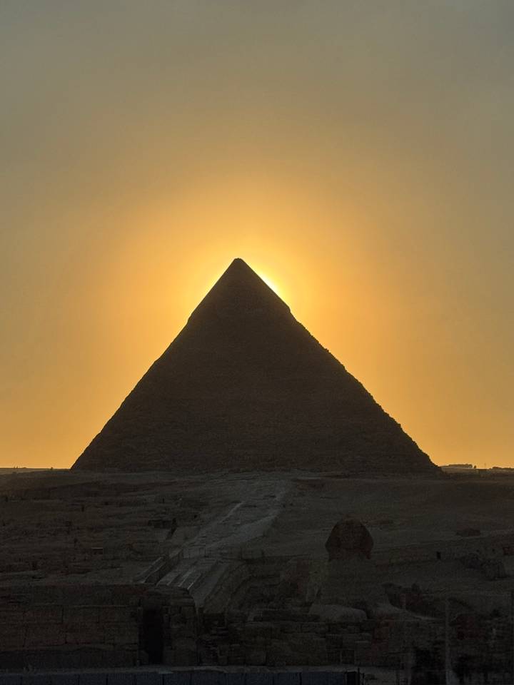 Silhouette of a pyramid perfectly aligned with the glowing golden sun behind at sunset.