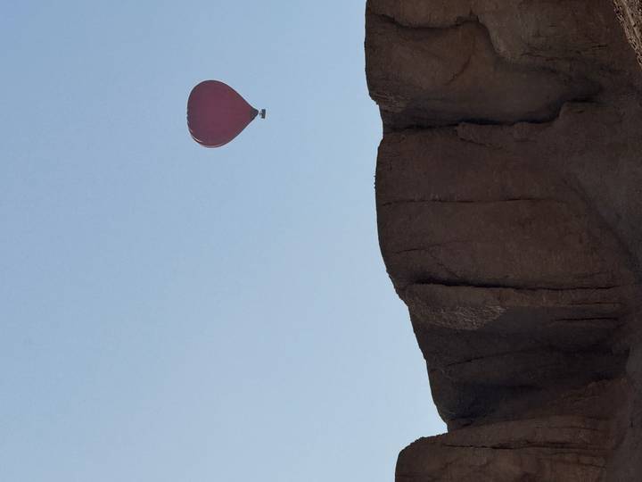 Red hot-air balloon floats high above a rugged desert cliff against a wide pale blue sky.