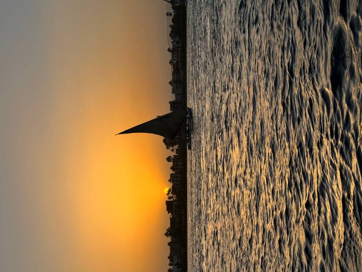 Traditional sailboat on rippling river water at golden sunset with urban skyline in silhouette.