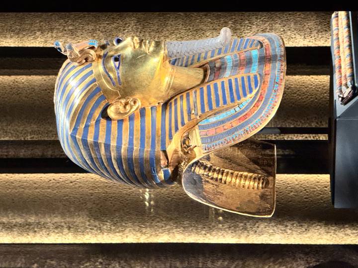 Gold and blue funerary mask of Tutankhamun displayed in a museum with dramatic lighting.