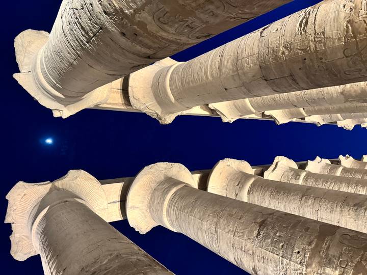 Illuminated stone columns of Luxor Temple reaching up to a deep blue night sky with a bright moon overhead
