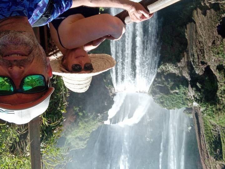 Selfie of two travellers in front of a powerful multi-tiered waterfall cascading through green cliffs.