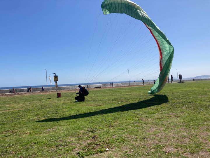 Paraglider canopy being laid out on a grassy seaside field while people stroll along the promenade