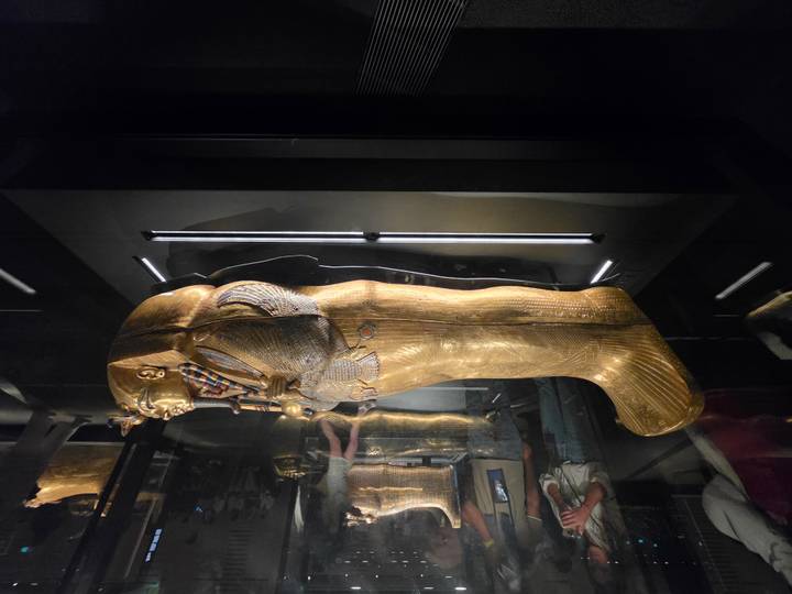 Golden pharaonic coffin encased in glass with onlookers in a darkened exhibit room.