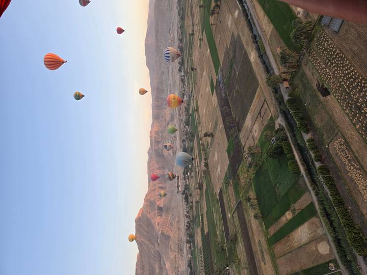 Dozens of colorful balloons drift above patchwork farmlands with desert hills in the background.