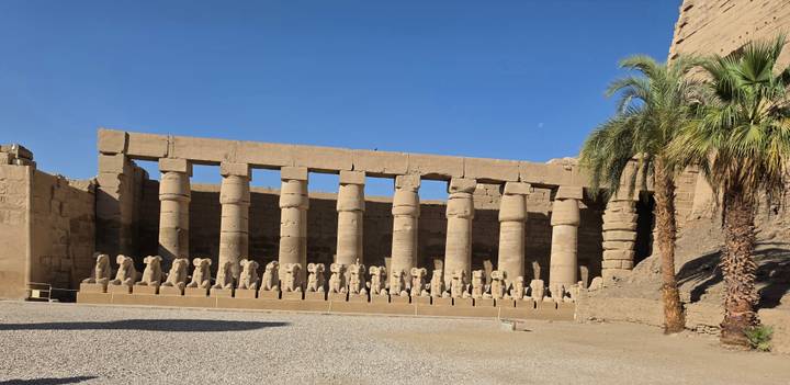 Row of colossal ram-headed sphinxes and towering columns inside Karnak Temple.