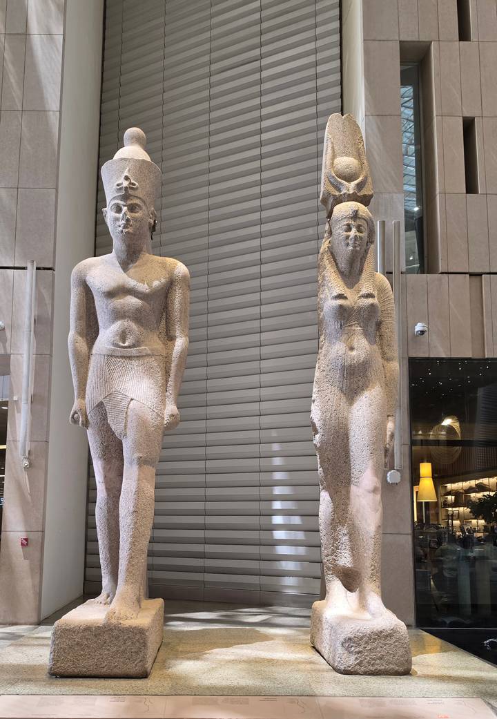 Two ancient stone statues displayed in a modern gallery with metal and glass surroundings.