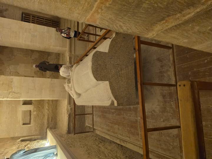 Stone granite sarcophagus displayed inside an ancient Egyptian tomb chamber with a few visitors observing.