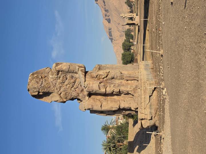 Sun-lit Colossus of Memnon statue towering against a blue sky with distant hills in Luxor.