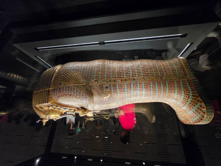 Colorful patterned sarcophagus exhibited in a museum glass case with visitors reflected around it.
