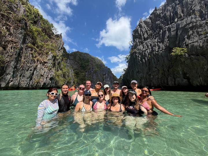 Group of swimmers pose in crystal-clear turquoise water flanked by towering limestone cliffs under a blue sky.