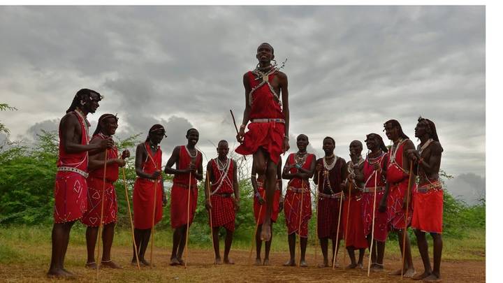 Maasai men dressed in red shukas performing a traditional jumping dance on dirt ground under cloudy skies.