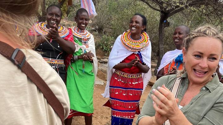 Travellers laughing and clapping alongside Maasai women in colourful traditional dress during cultural visit.